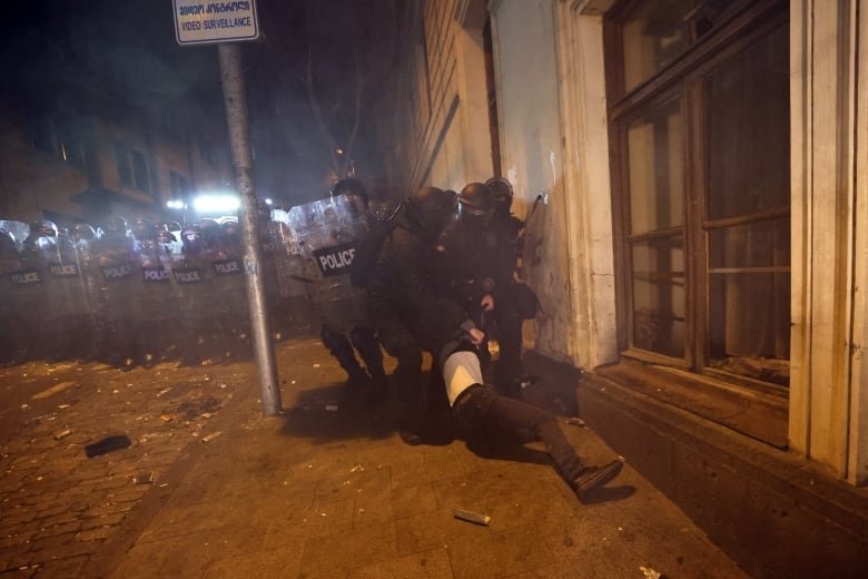 Several officers in riot gear are shown on a sidewalk against a building, dragging a person.