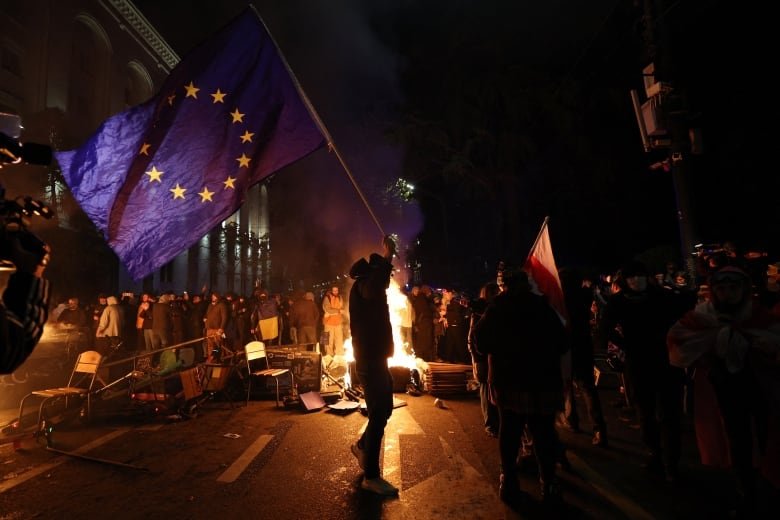 A silhouetted person wearing a hat, holds up a large flag on a city street at night, with a fire burning behind him and several onlookers.