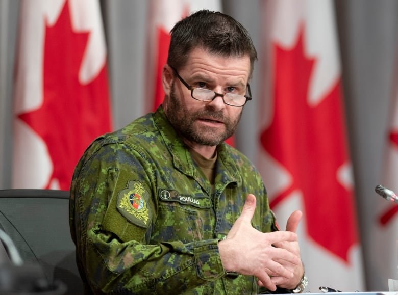 A man in a green camouflage uniform speaks in front of a line of Canadian flags.