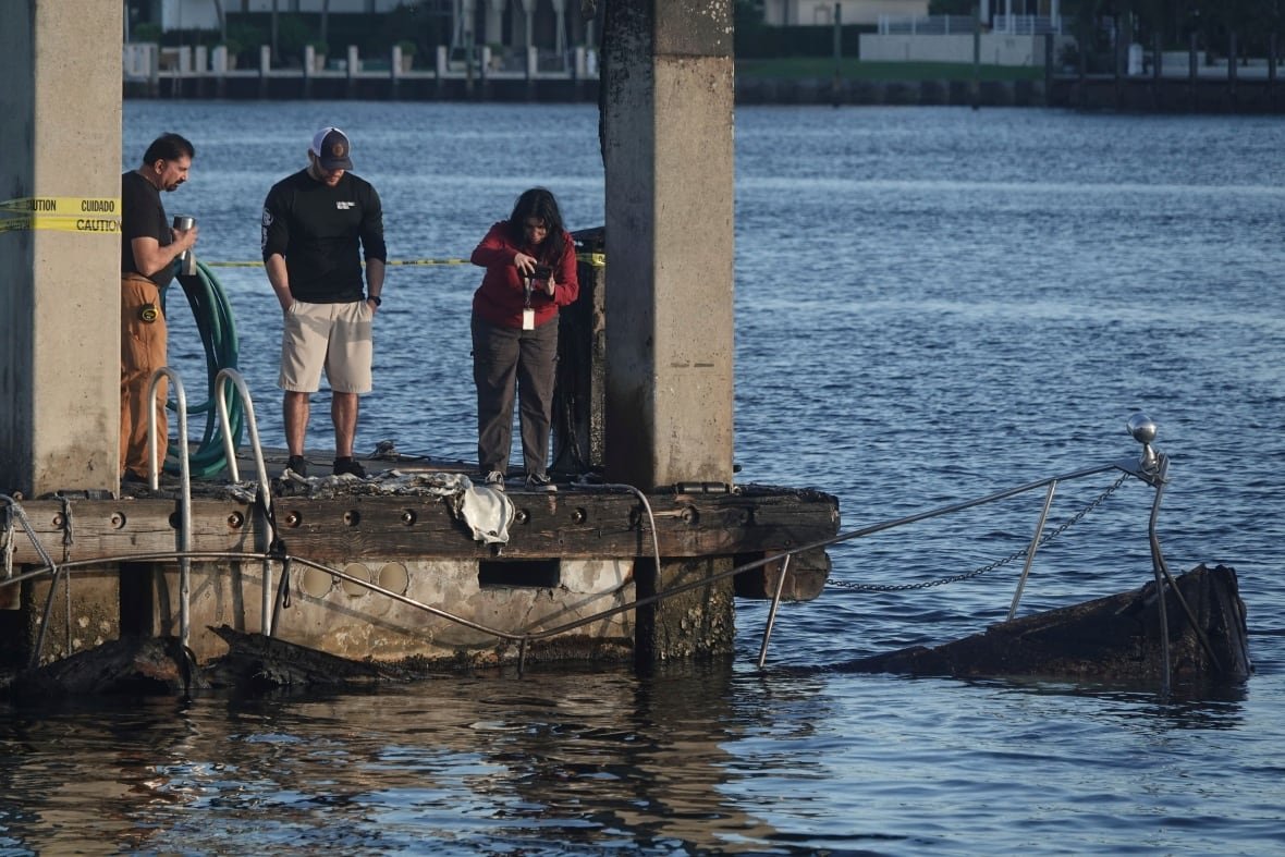 boat-explosion-florida-1 People standing on the edge of a pier survey the charred remains of a boat in the water.