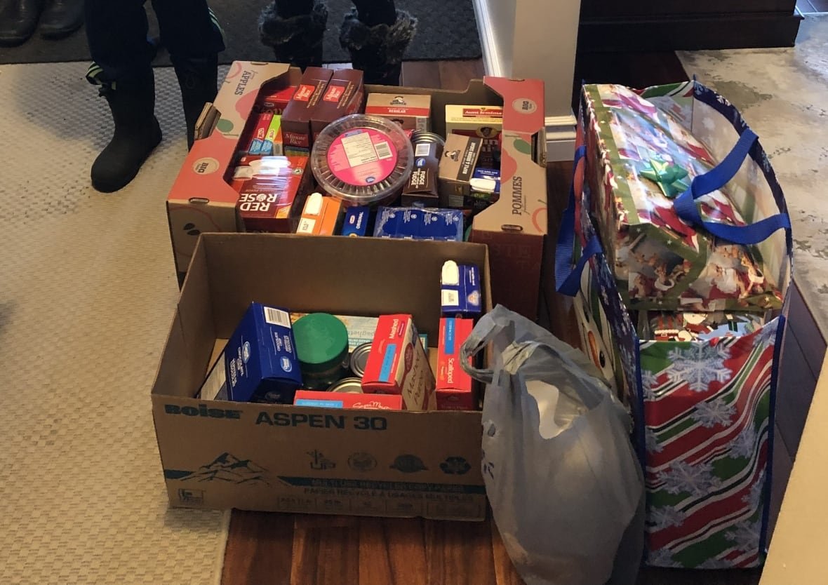 christmas-hamper Two cardboard boxes filled with non-perishable food and a large bag with boxed presents sit on the floor.