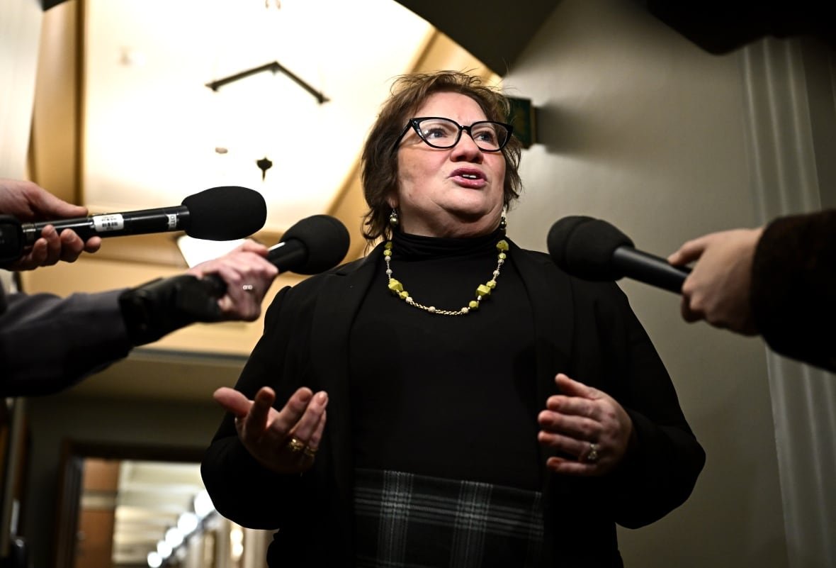 A woman wearing a black turtle neck and gold necklace speaks as reporters hold microphones.