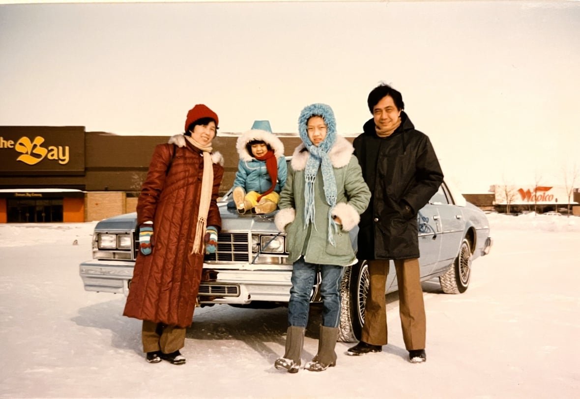 linda-trinh-1 A smiling family stands next to a car while a toddler in a blue snow jacket sits on the hood. They’re in a parking lot for The Bay and it’s covered in snow.
