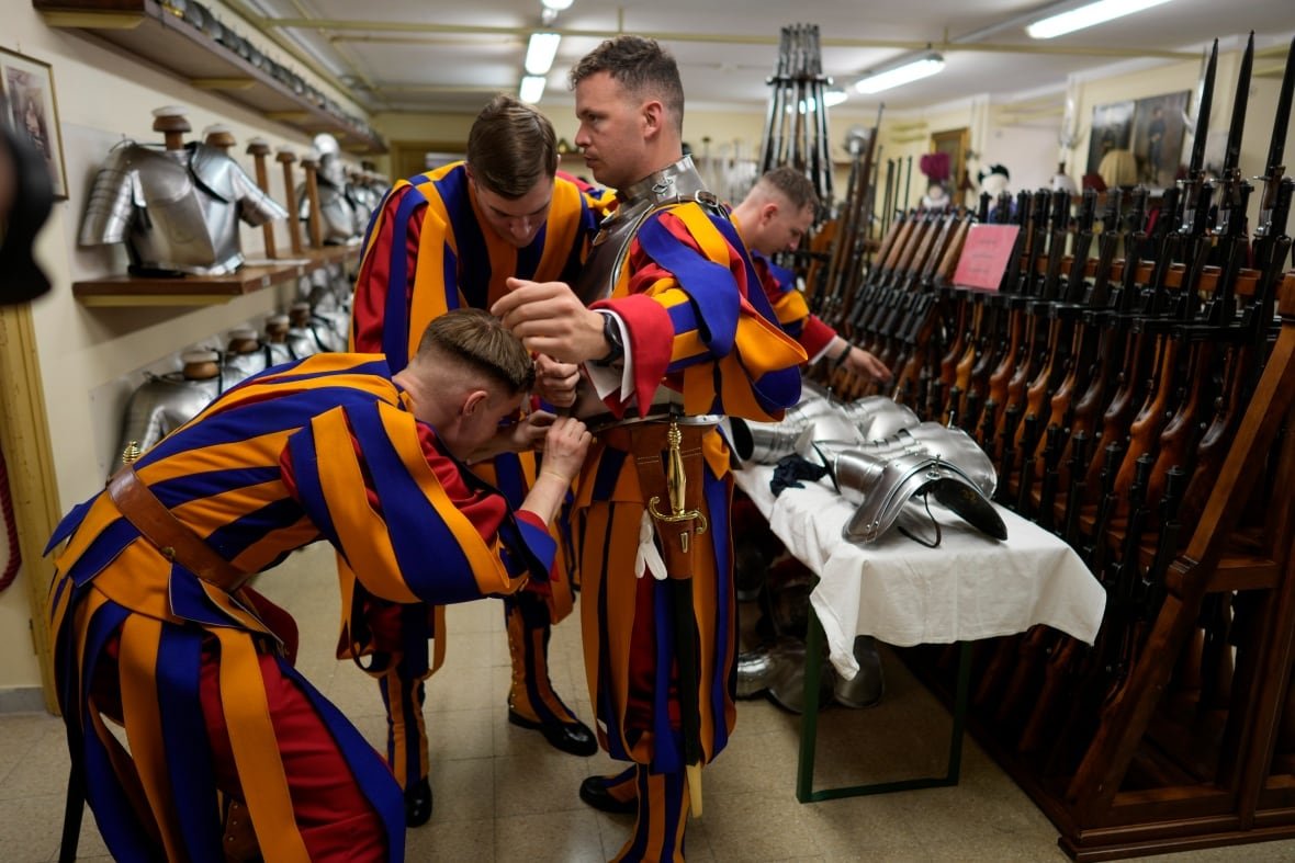 vatican-swiss-guards-1 Two men in striped coloured outfits assist a third man with his outfit.