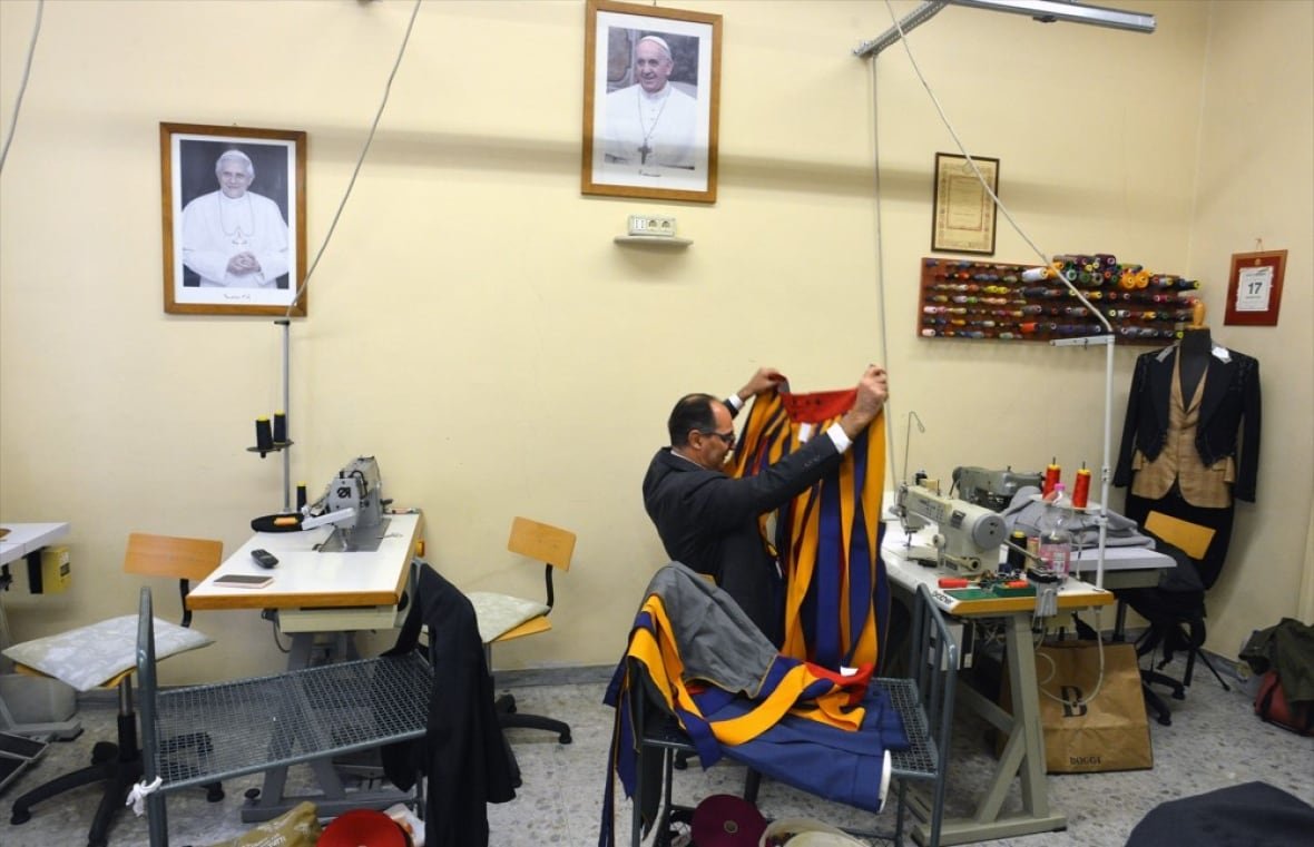 vatican-tailor-3 A man seated at a sewing machine holds up a yellow and blue striped garment.