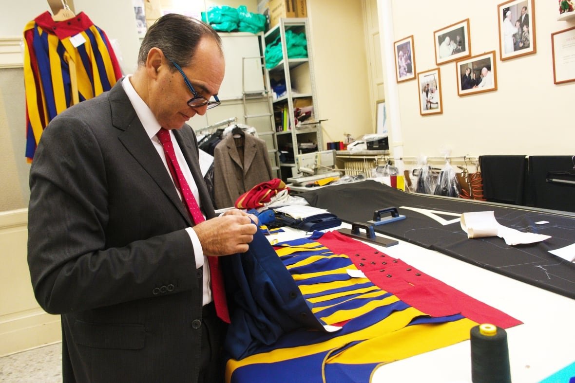 vatican-tailor-esma-cakir A man in a grey suit and red tie stitches material while standing in front of a work bench covered in coloured material.
