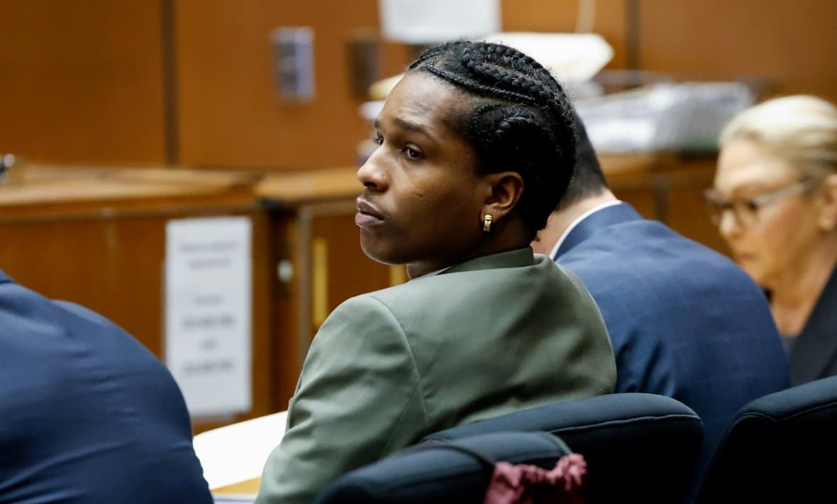 A man in a suit sits back in a chair in a courtroom, looking off to the left behind the back of the man sitting next to him.