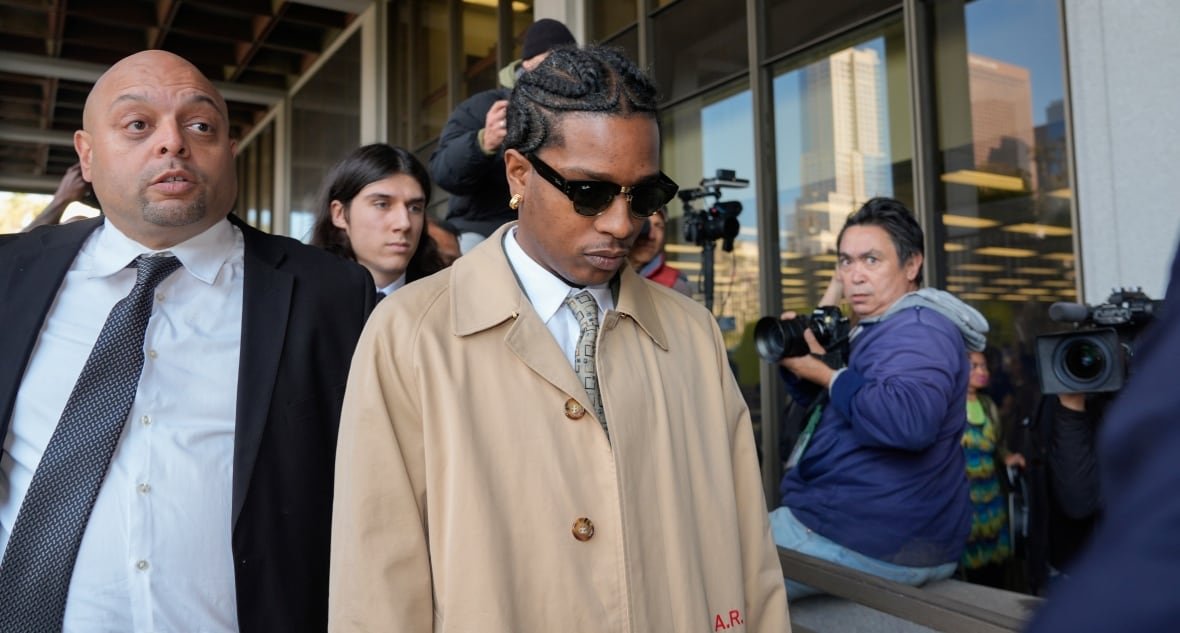 A man in a beige coat and dark sunglasses walks with his head tipped down. He is surrounded by other people, most carrying cameras.