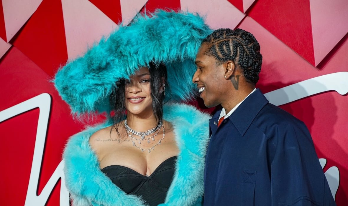 A couple are shown on a red carpet, posing in front of a red backdrop. The woman is wearing a massive, cyan feathered hat that matches the bright cyan fluffy coat she is wearing over a black dress. She is smiling at the camera, while the man smiles at her.
