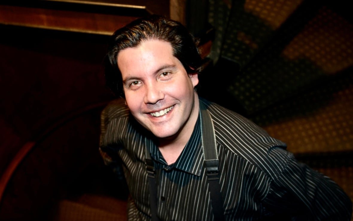 curtis-brandell-1 A man with black hair, a dress shirt and suspenders looks up at the camera with a smile.