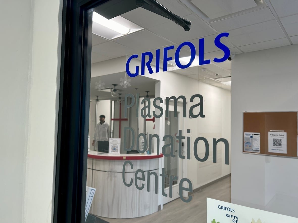 grifols-plasma-donation-centre-1 A glass door with the words Grifols Plasma Donation Centre, and a blurry man in the background walking toward a reception desk.