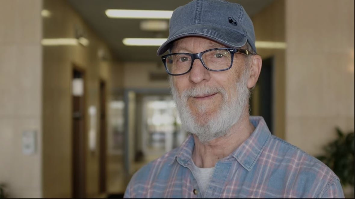 jean-claude-basque-1 A man with a white beard, plaid shirt and blue hat, smiles at the camera with a hallway behind him.