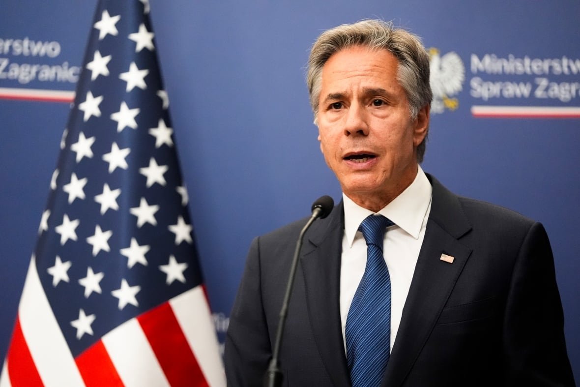 poland-us-blinken-1 A gray-haired man in a dark suit and blue tie speaks into a microphone. An American flag is behind him.