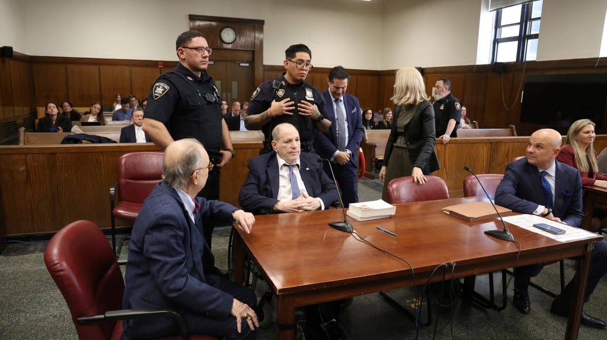 sexual-misconduct-harvey-weinstein-4 An old man sits at a brown desk in a courtroom, with people standing and sitting around him, including two police officers standing right behind him.