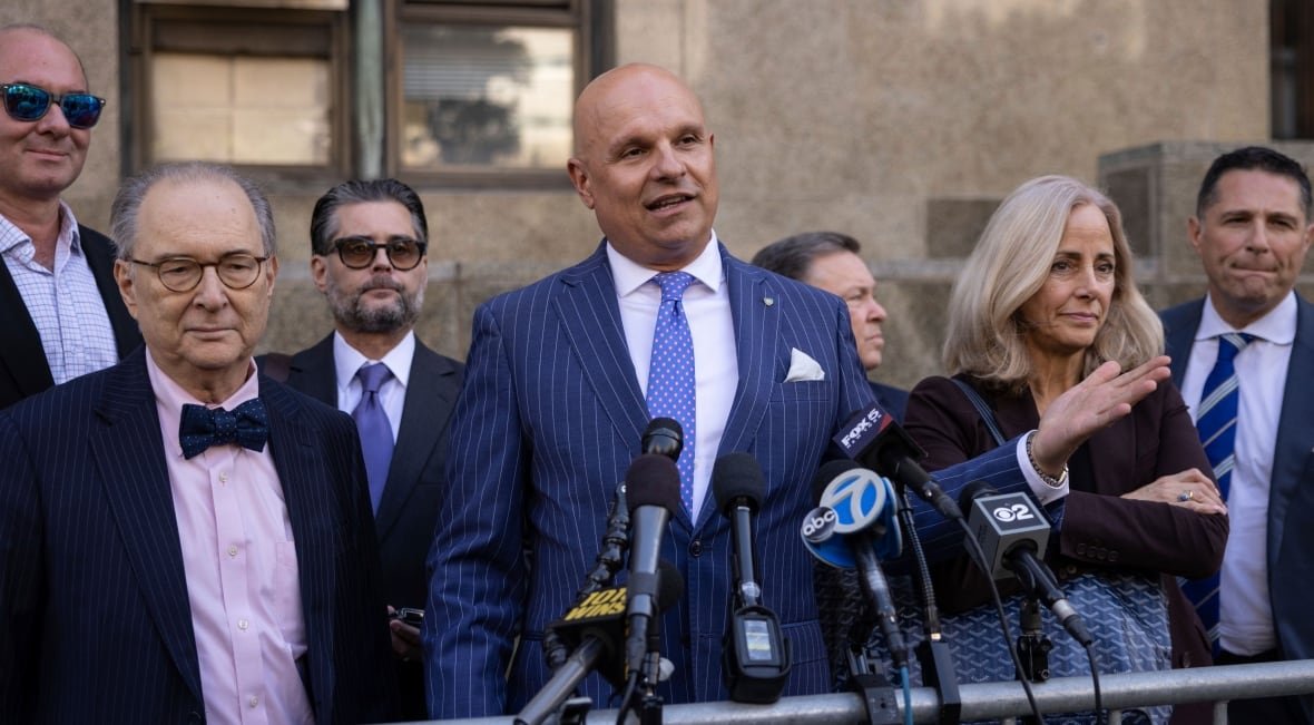 sexual-misconduct-harvey-weinstein-5 A man in a blue suit stands outside in front of a fence and a row of microphones, flanked by a bunch of other people.
