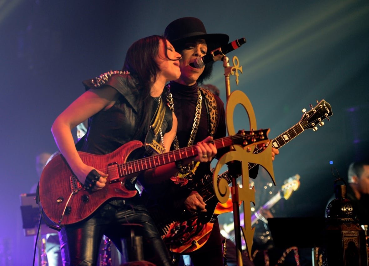 477471851 A woman playing a guitar and a man holding a guitar stand side by side singing at a microphone on a concert stage.