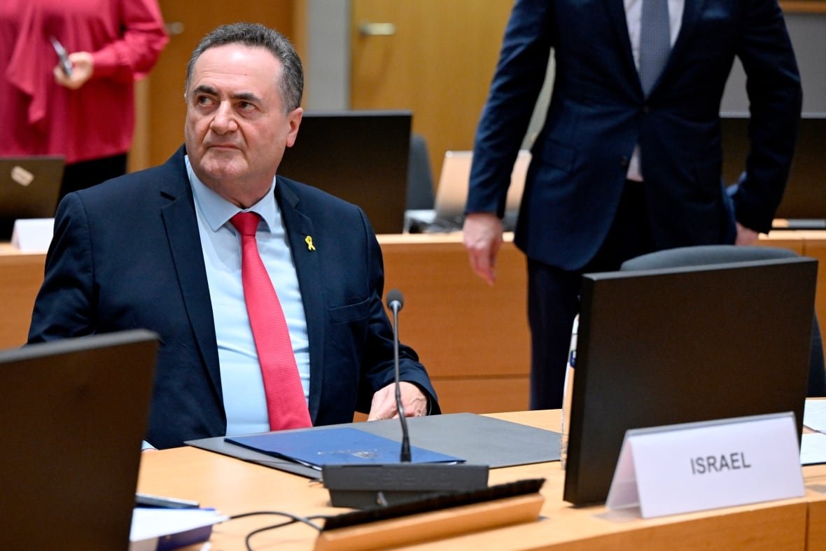 belgium-eu-foreign-ministers-1 A man, wearing a blue blazer and red tie and yellow ribbon on his lapel, sits in a meeting room at a desk behind a microphone.