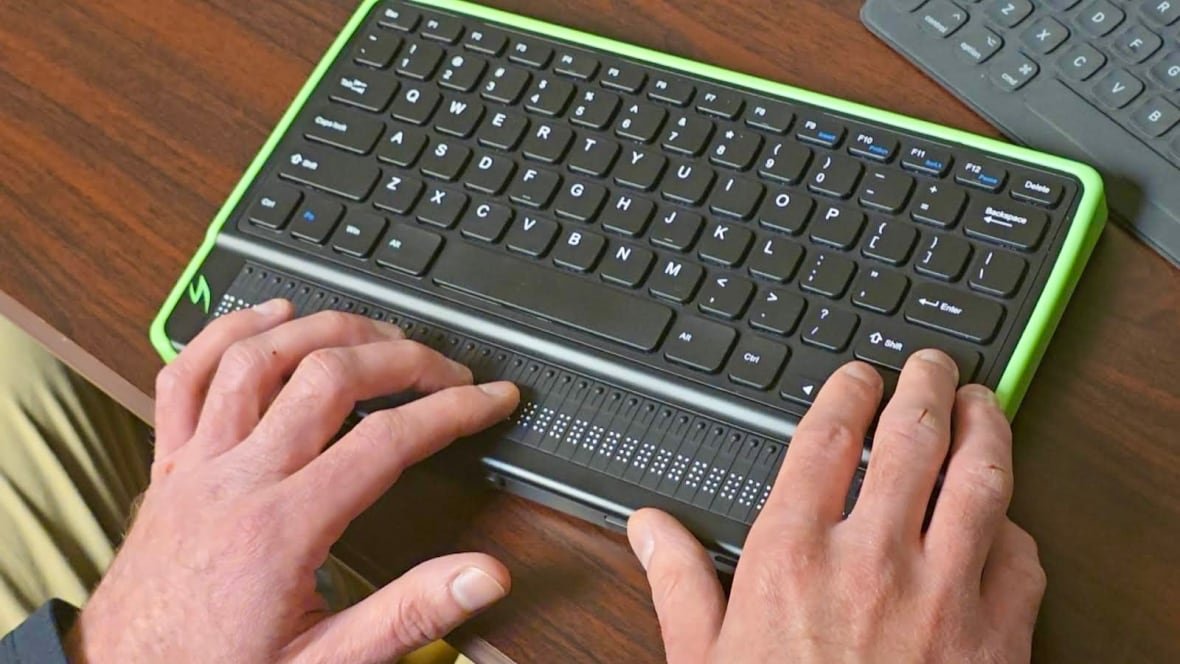 braille-keyboard Two hands touching a braille keyboard sitting on a wooden desk.
