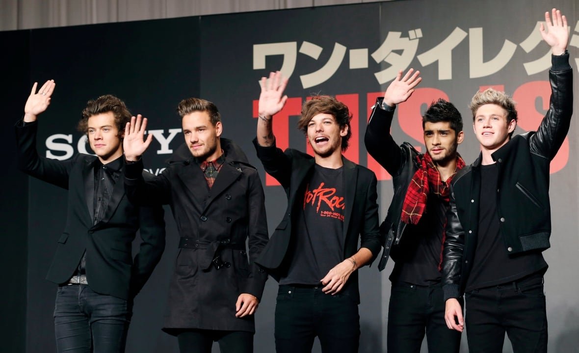 britain-one-direction Five young men wave on a red carpet, most wearing all black outfits. There is a black backdrop behind them with Japanese on it.