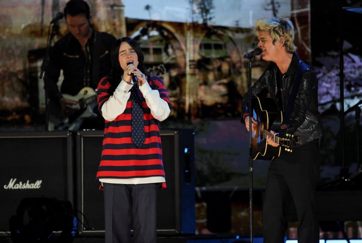 california-wildfires-concerts-1 A woman wearing a black and red striped top with a tie sings on a stage with a man playing a guitar.