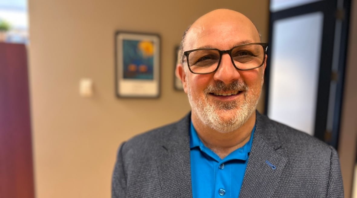 charles-levasseur A man with short hair, short beard and black rimmed glasses smiles at the camera, with a grey suit jacket over a blue shirt.