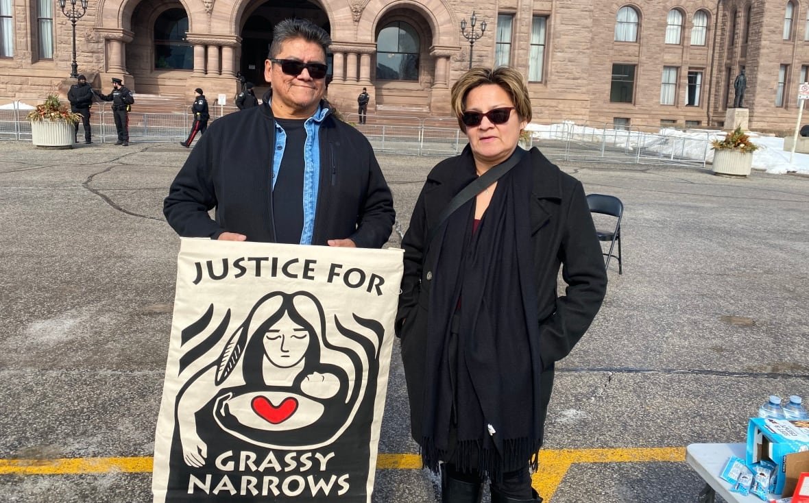 coun-aaron-kokopenace-and-chief-sherry-ackabee Two people are seen standing outside a large building, holding a sign that says "Justice for Grassy Narrows."