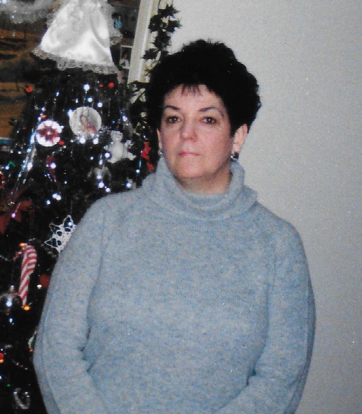 covid-anniversary-deaths-20250305-5 Patricia West is shown in an undated photo standing in a light blue sweater in front of a Christmas tree.