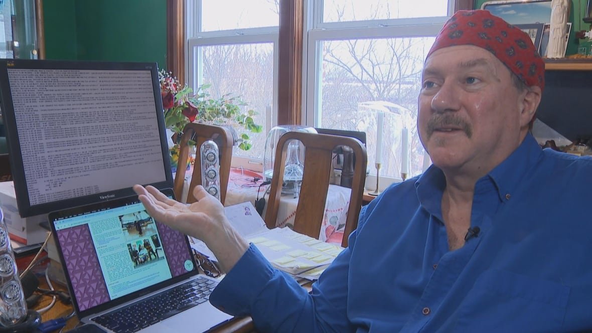 daniel-mackay An older man sits in front of a desk. At the desk, there's a computer with a white screen and black text with code. There is also a laptop, which shows an article.