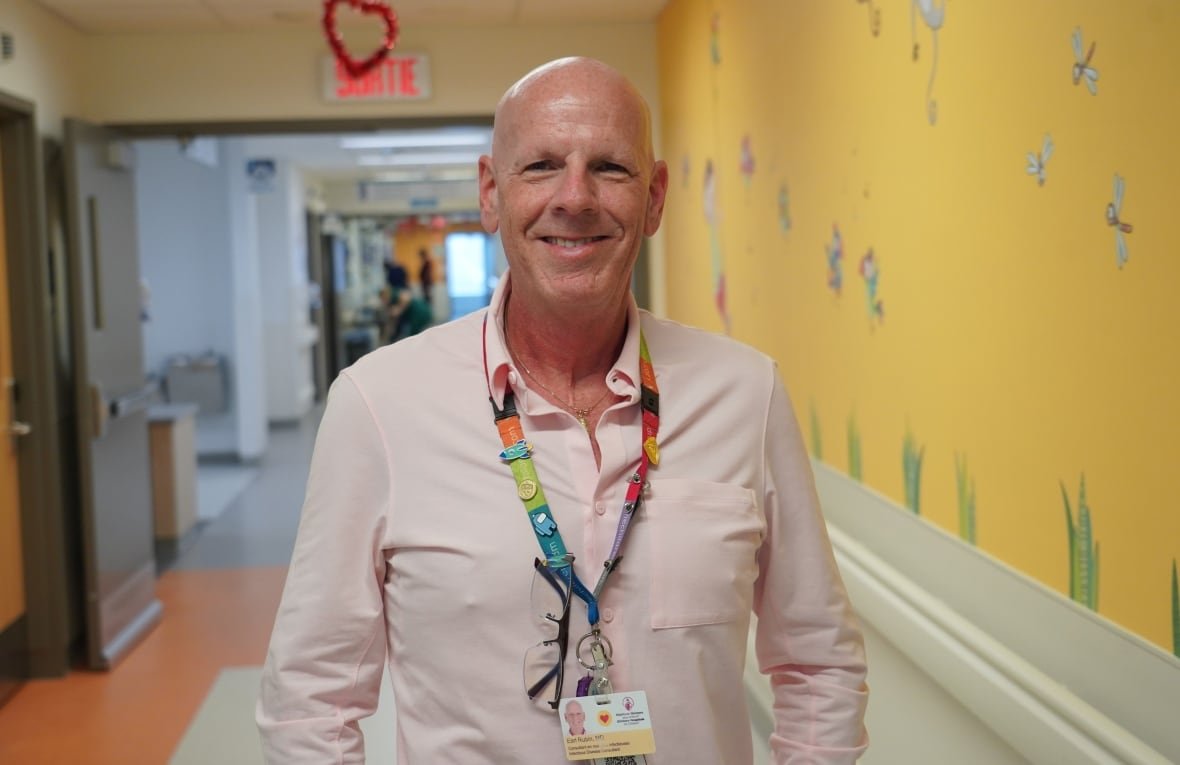 dr-earl-rubin A bald man wearing a light coloured shirt and multicoloured lanyard stands smiling in a corridor with a yellow painted wall with grass and insect decals.