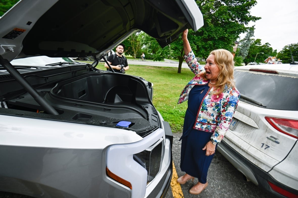 electric-vehicles-federal-government-policy-2023 A politician opens her mouth in surprise as she opens the front hood of an electric vehicle.