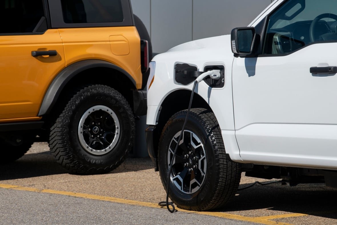 ford-electric-vehicles The front end of a white pickup truck is pictured with a charging cable plugged in above the driver's side front tire.