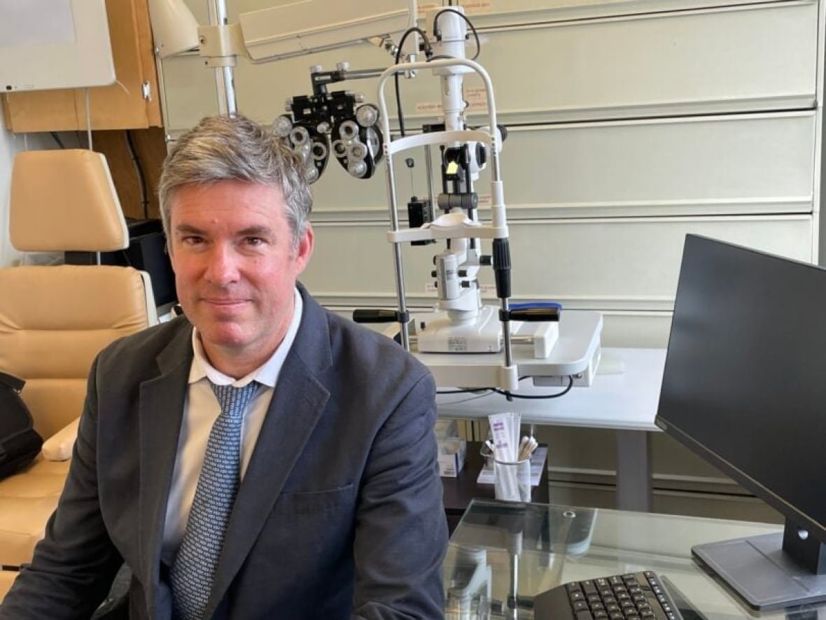 greg-moloney A grey-haired man in a suit sits next to a computer in front of an eye exam chair and machine.