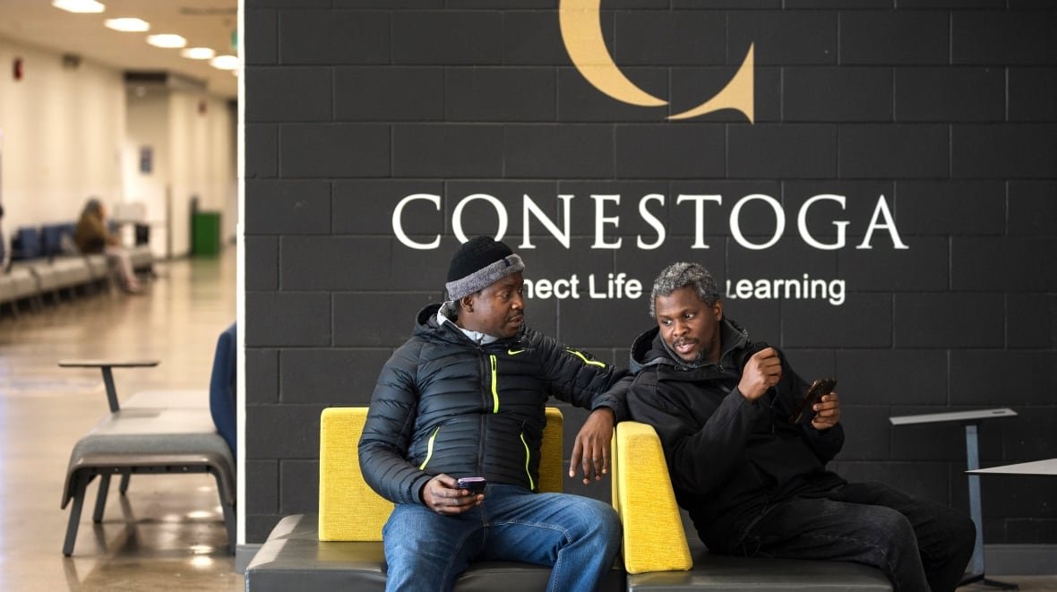 international-students-20240430 Two students in conversation, seated in front of a wall with the Conestoga College logo.