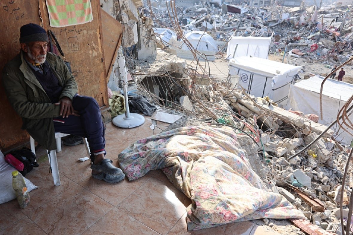 jabalia-1 An older man sits on a chair in a destroyed house. Outside there are white tents.