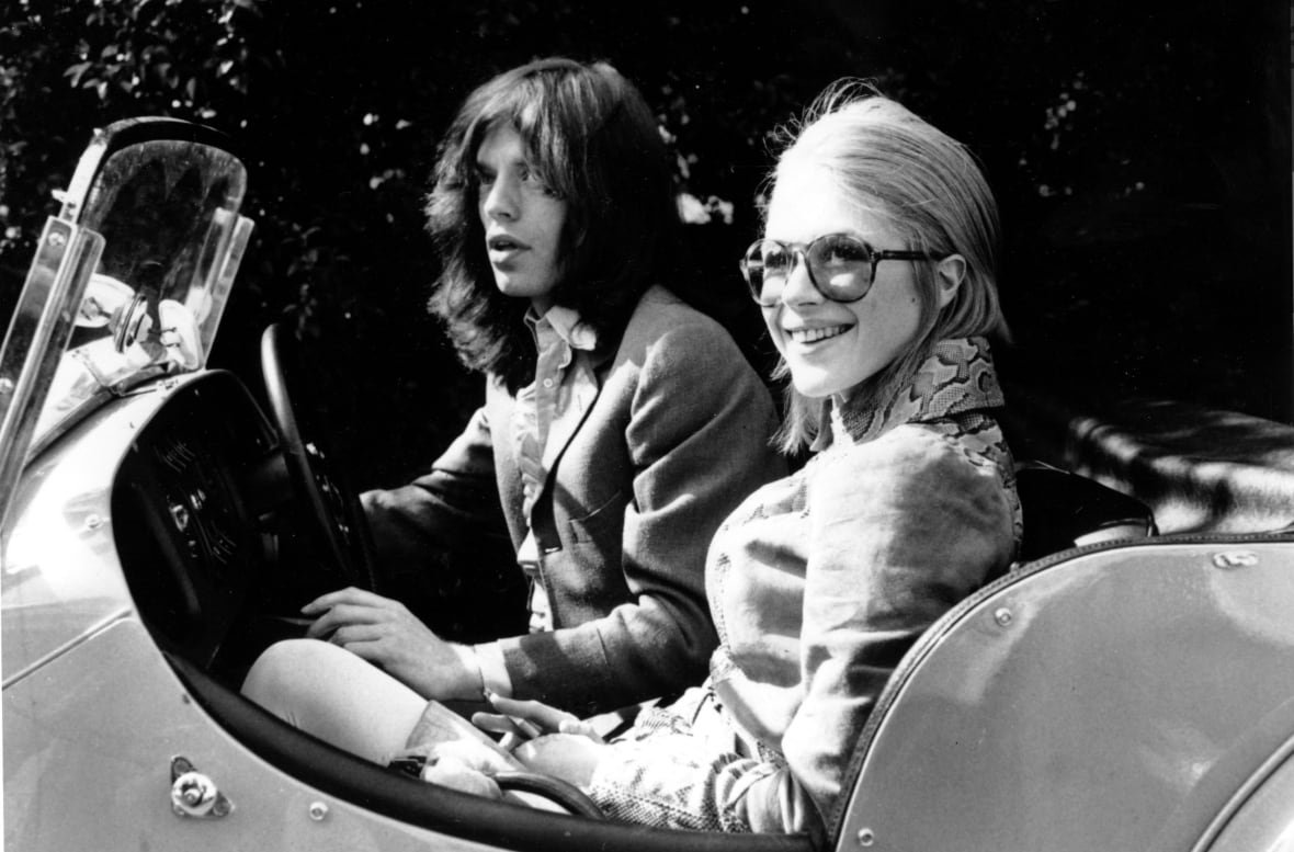 jagger-faithfull In this black and white photo, a couple sit in a car, looking to the side at the photographer standing next to the car. The woman is closer to the camera and is smiling. The man has long hair.