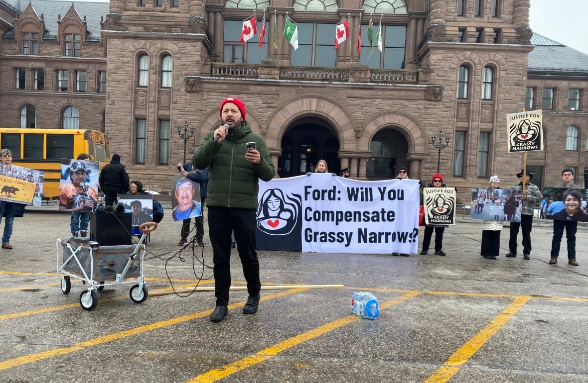 jeffrey-ansloos A person is seen standing in front of a group of people outside who are holding signs. A large banner behind the person standing says "Ford: will you compensate Grassy Narrows?"