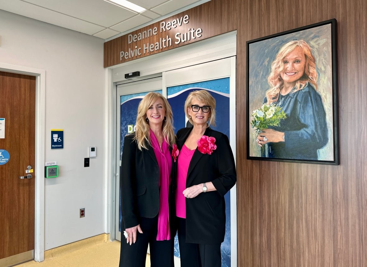 Two women dressed smartly in black and hot pink stand in front of a doorway to a hospital next to a portrait of a woman.