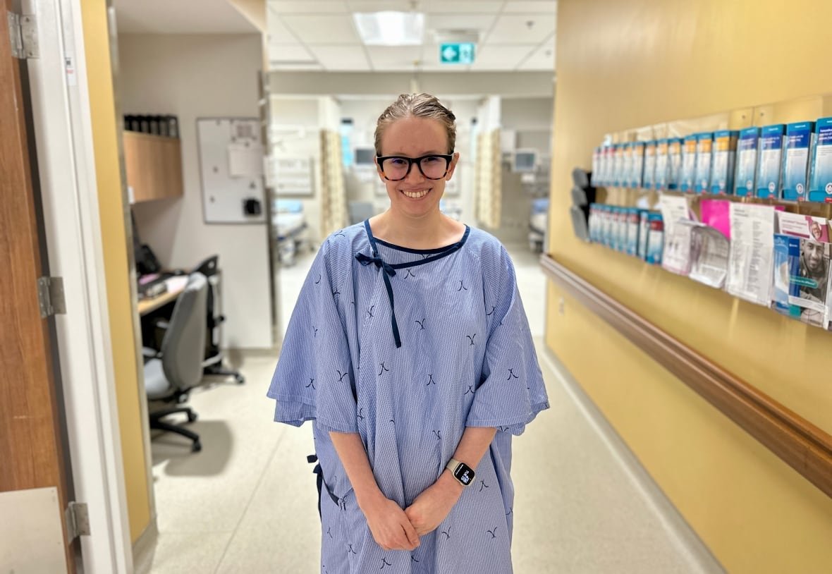 A woman wearing glasses in a blue hospital gown stands in a hospital hallway and smiles.