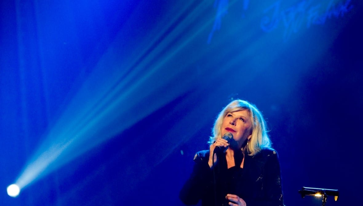 marianne-faithfull-obit-1 A woman sings into a microphone in front of a blue backdrop. A spray of stage lights shines from the bottom left corner of the image up diagonally over her head.