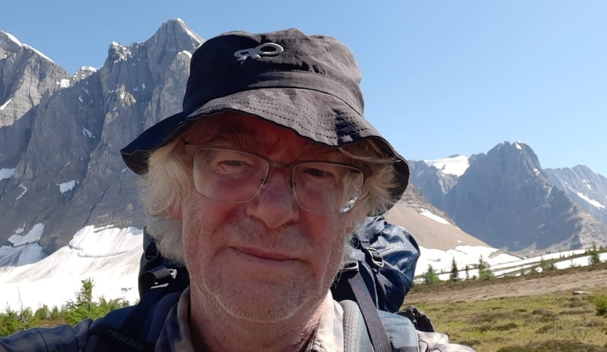 nigel-bankes A man wearing a hat looks at the camera with a backdrop of mountains pictured behind him.