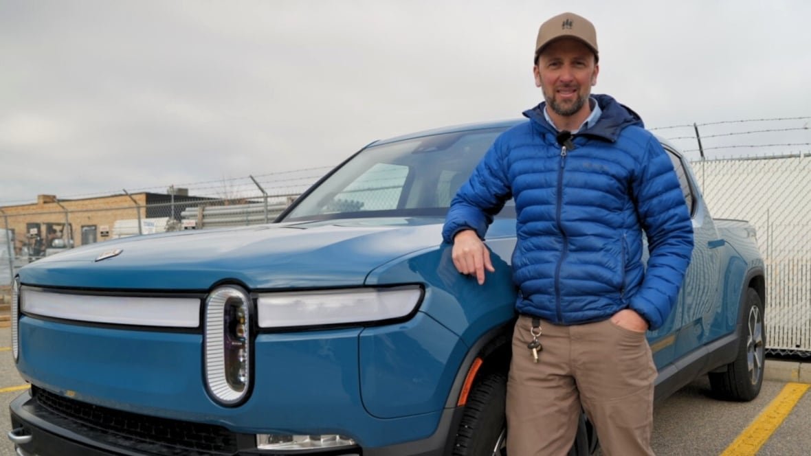 richard-wilton A man in a blue jacket stands beside an electric Rivian truck which is also blue.