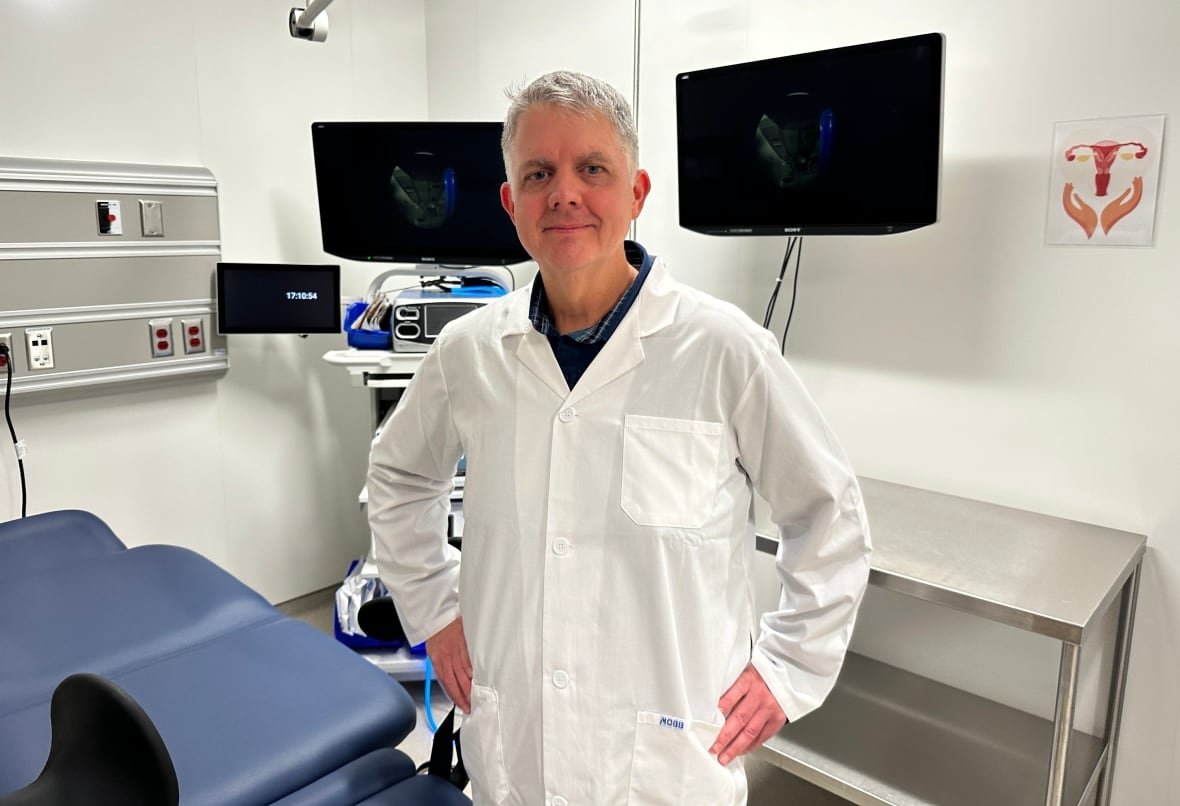 A male doctor in a lab coat stands and smiles with his hands on his hips.