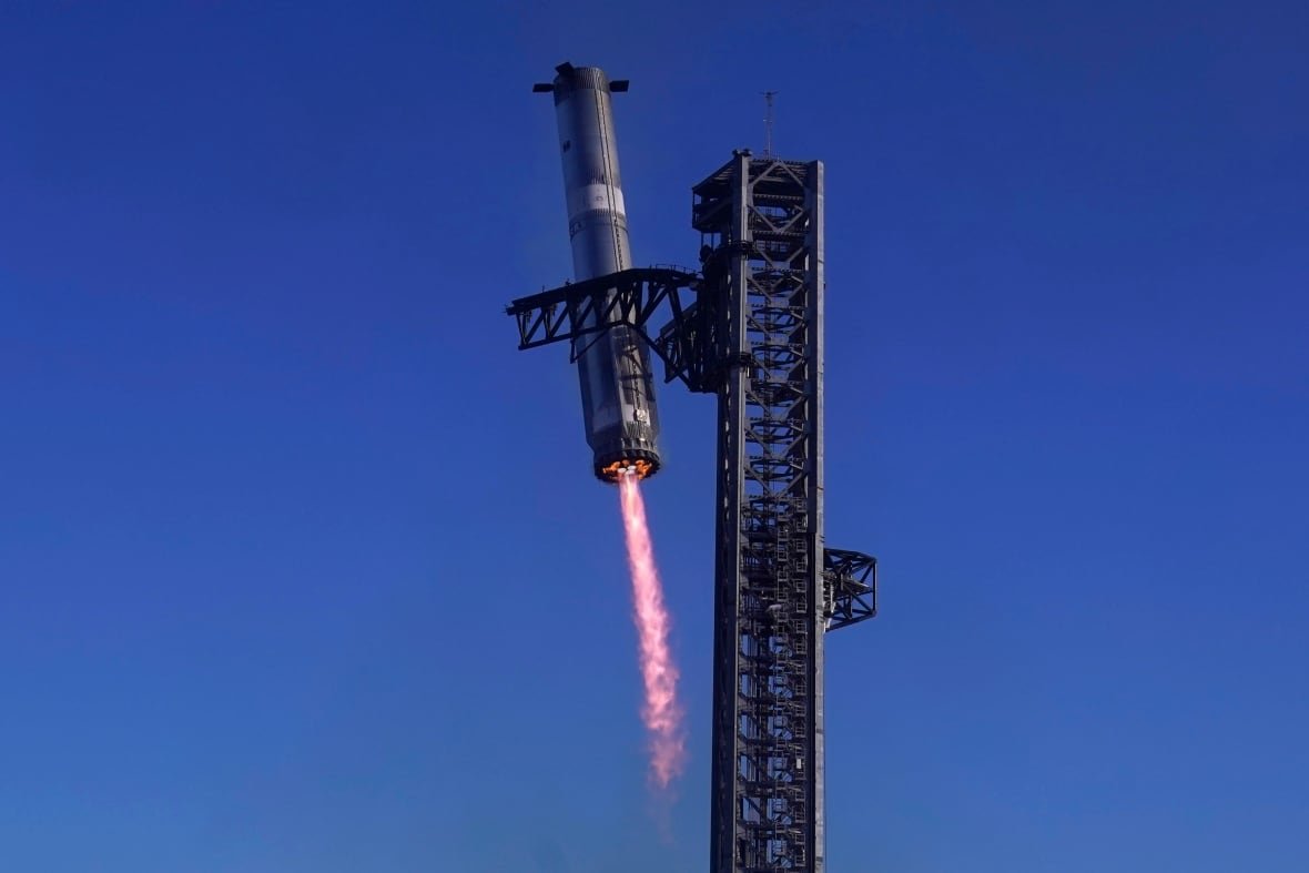 spacex-starship-launch-1 A rocket, a gout of flame coming from its rear, is grabbed by mechanical arms on a large tower.