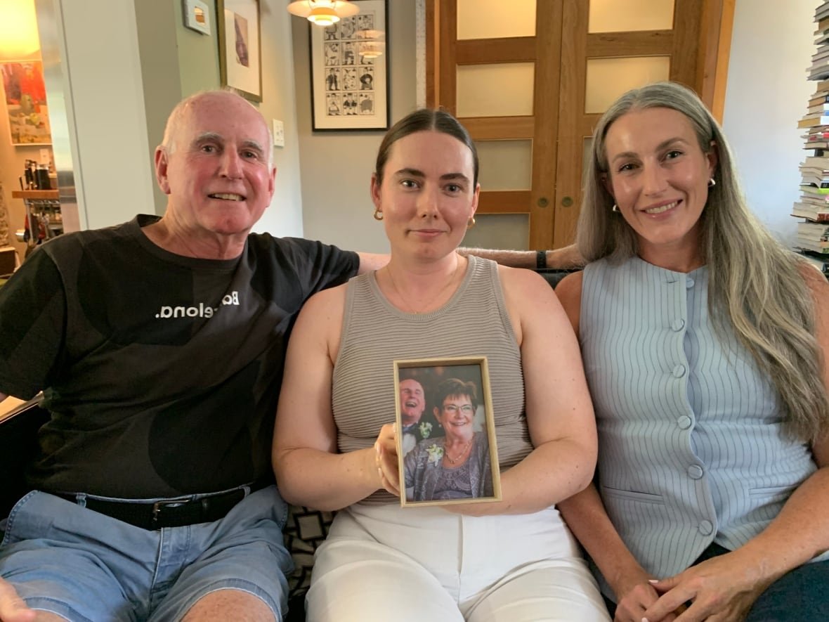 terry-mccarthy-molly-mccarthy-and-tara-kerr-1 A bald man sits next to two younger women on a sofa. The woman in the middle is holding a portrait of the older man and his wife.