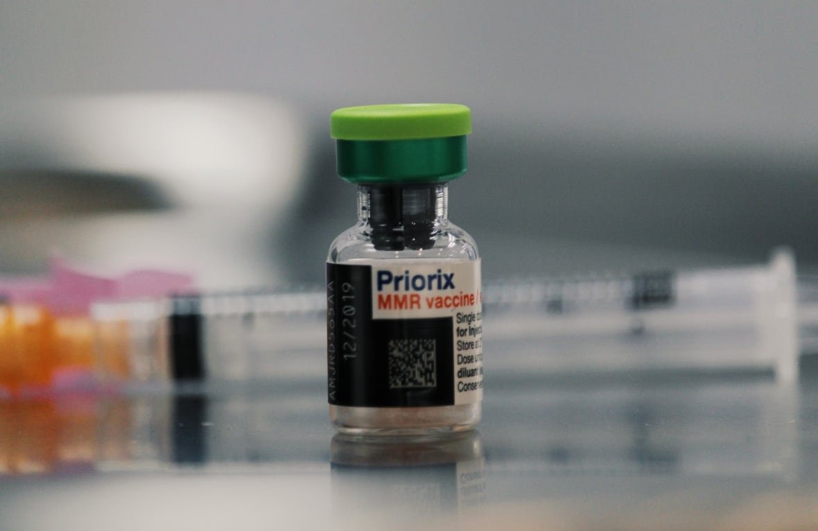 A vial of measles, mumps and rubella vaccine with out of focus syringes behind it.