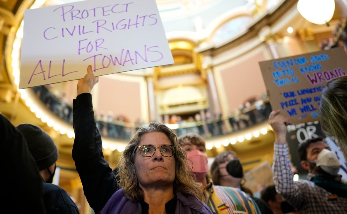 transgender-civil-rights-2 A person with a determined expression holds a sign up that says "Protect civil rights for all Iowans." A crowd is visible behind them, with more people holding signs.