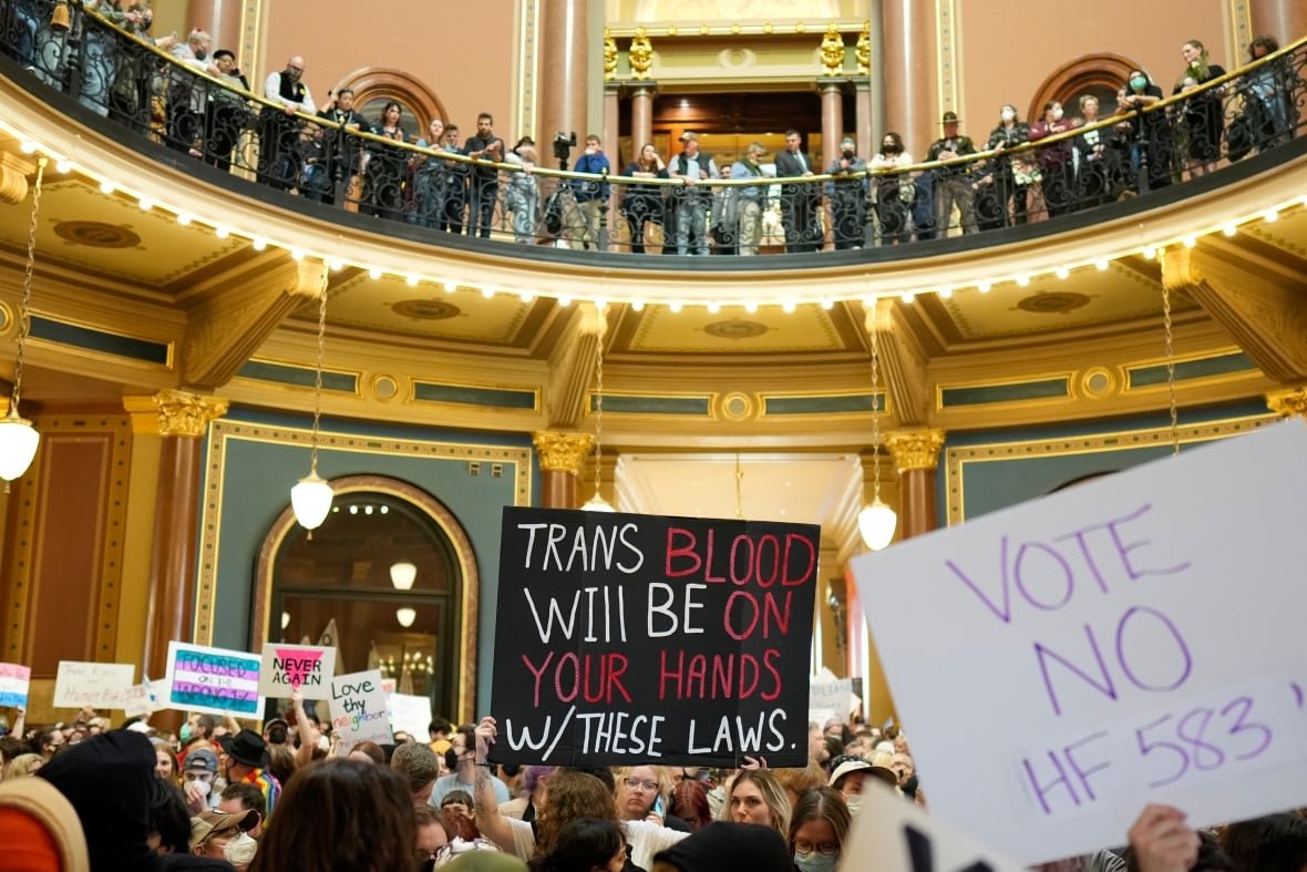 transgender-civil-rights-4 A crowd of people is shown in a large, fancy room, with more people lined up on a balcony level above. The focus is centred on one sign held above someone's head in the middle of the room which says "Trans blood will be on your hands w/ these laws". Other people hold similar signs expressing support for the transgender community.