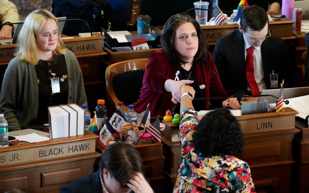 transgender-civil-rights-5 A woman seated behind a long wooden desk holds the hand of another person who is sitting in the row in front of her and has turned to extend their hand. Several other people stand or sit in the rows around, many looking upset or stressed.