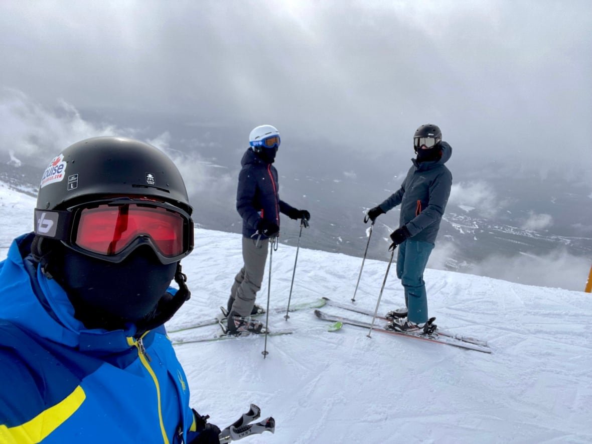 dejan-ratkov-1 Three people in snowsuits and ski goggles standing on a snowy ski hill.