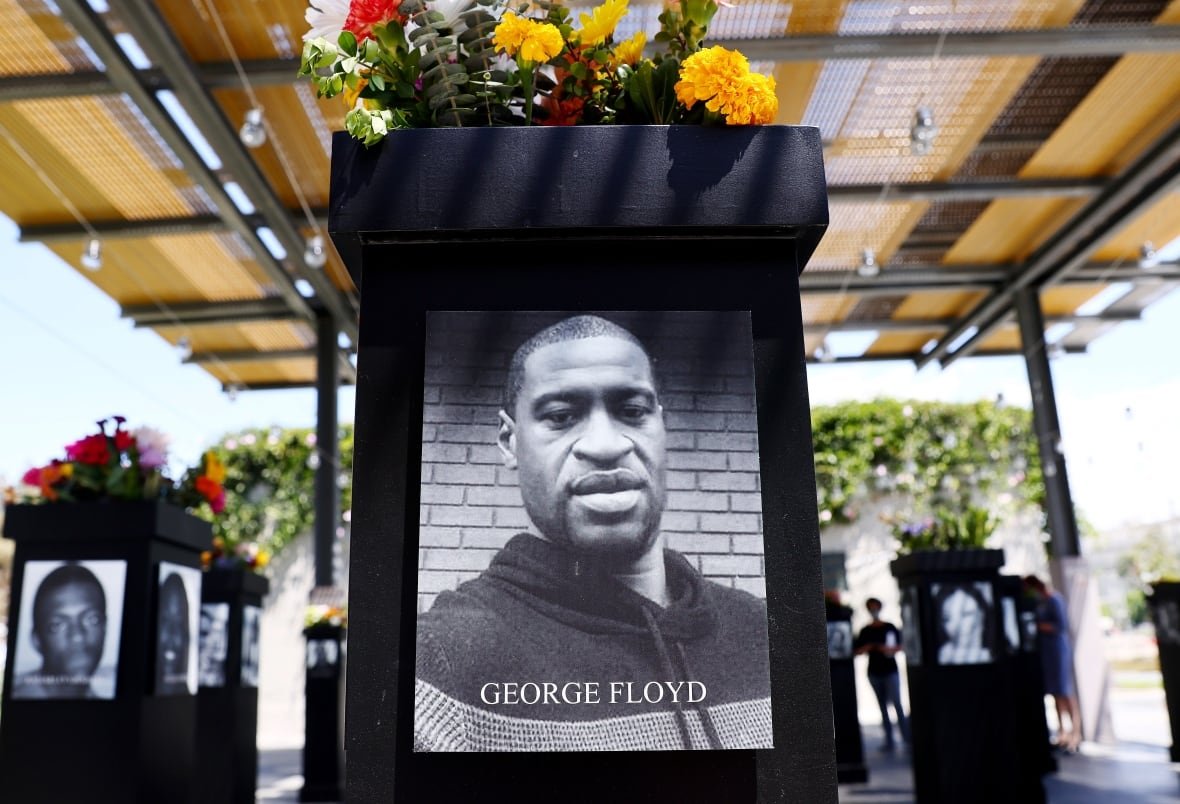 1329772991 Black and white phot of a Black man in a hoodie displayed on a monument covered in flowers with the words: "George Floyd"